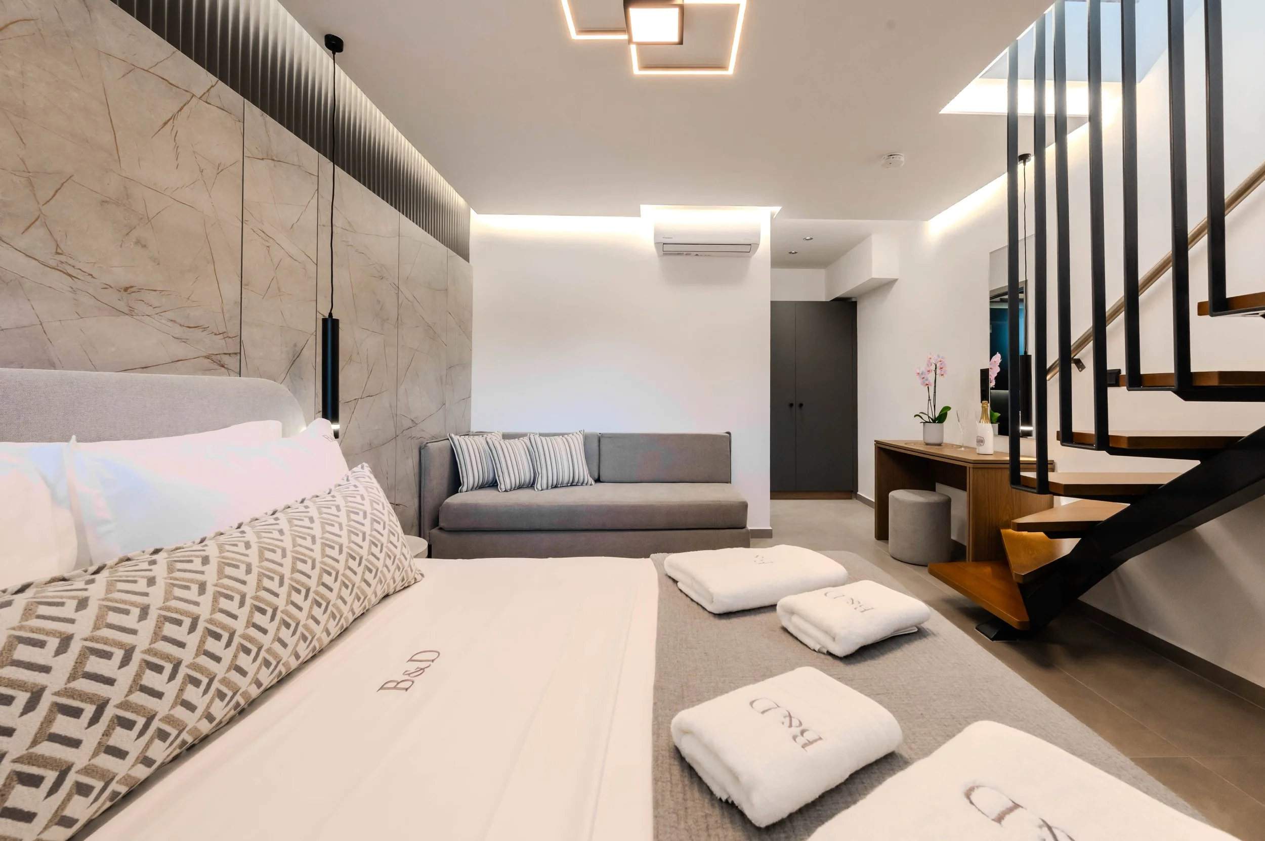 Modern hotel room with a bed, folded towels, a gray sofa with pillow, wall-mounted lamp, wooden desk with potted plants, staircase with black railing, and minimalist decor.