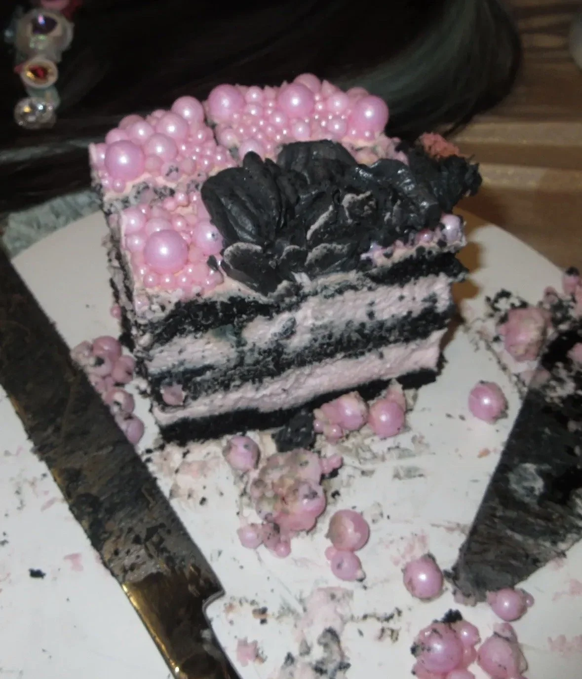 A slice of cake with pink and black decorations, including pink pearls, on a white cake board with crumbs and embellishments surrounding it.