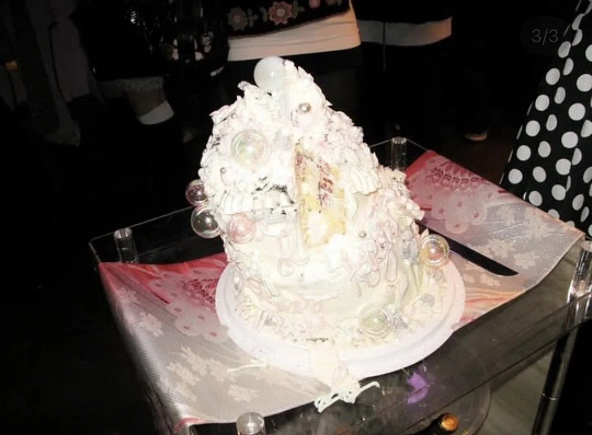 A large cake with white and pastel-colored frosting decorated with various ornaments, displayed on a glass table with a pink and white tablecloth. People stand around the table.