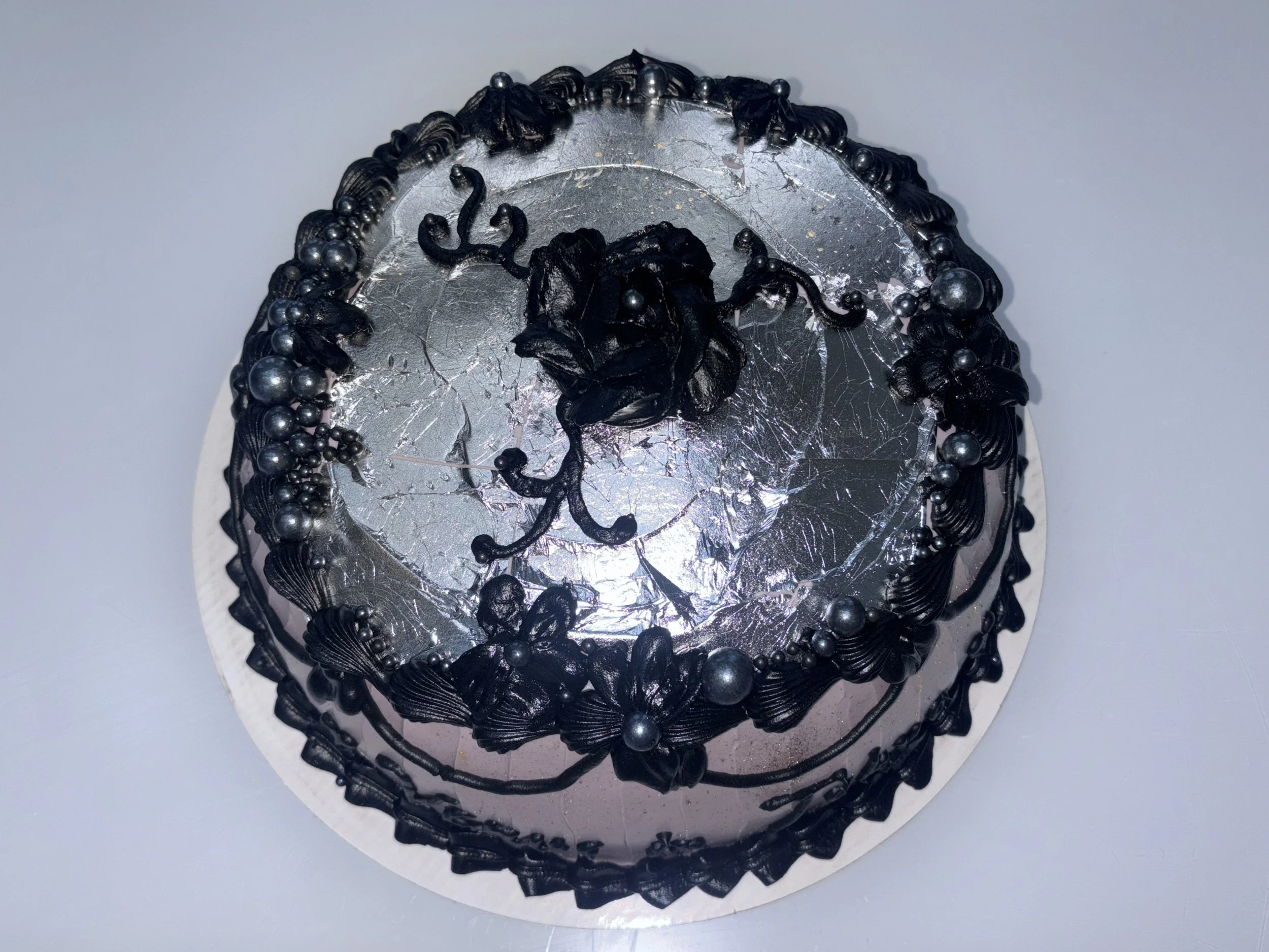Black decorated cake with floral and pearl patterns on black frosting, topped with a black flower, and silver foil