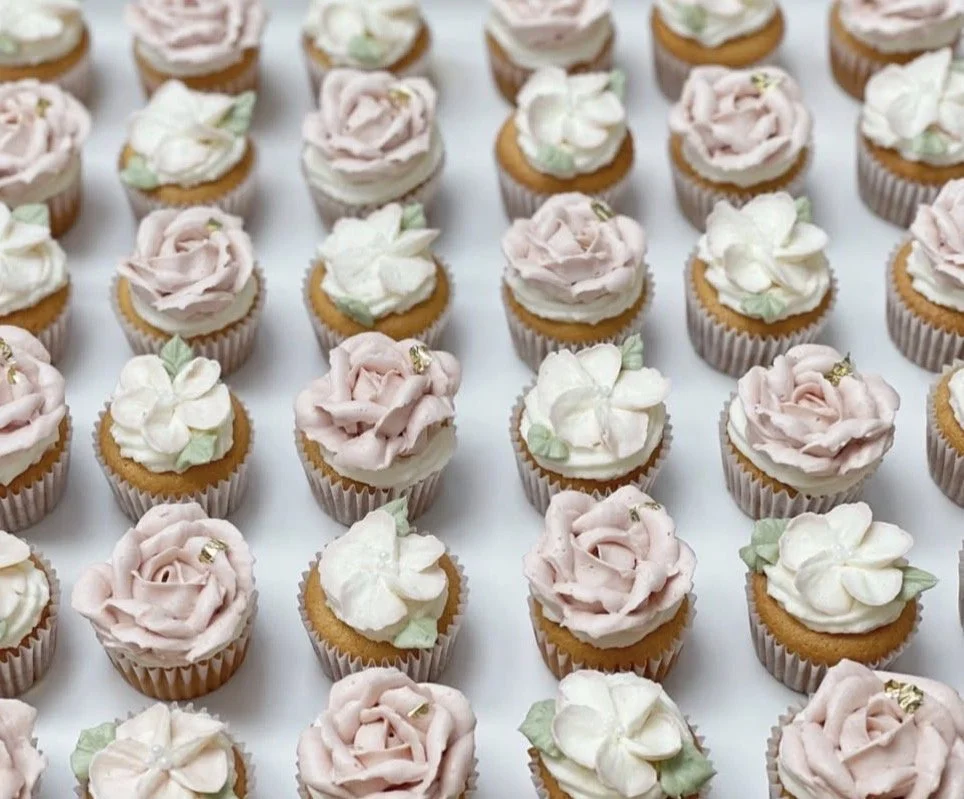 Mini cupcakes decorated with pink and white frosting designed to resemble roses, with small green leaf accents and gold sprinkles, arranged in neat rows on a white surface.