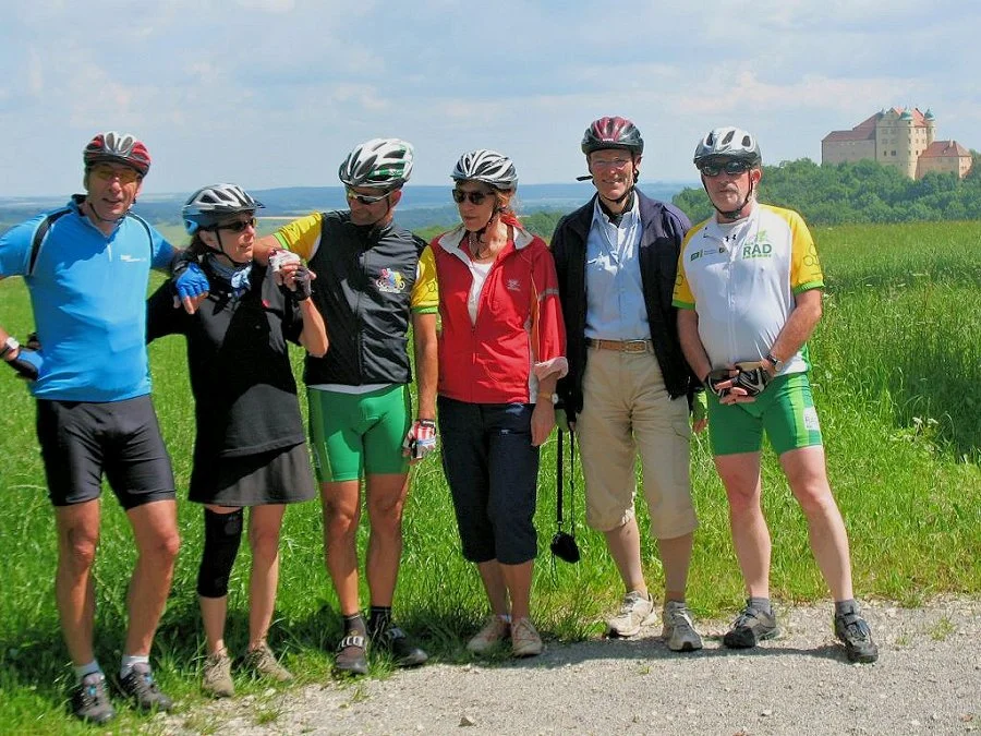 Sechs Radfahrer stehen zusammen auf einer Wiese am Tag, einige tragen Helme und Radkleidung, im Hintergrund eine Burg auf einem Hügel.
