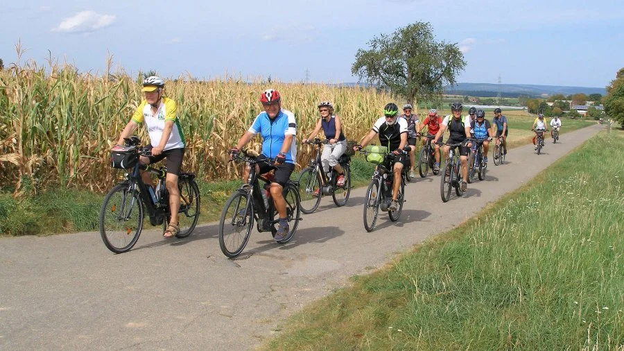 Eine Gruppe von Menschen fährt mit Fahrrädern auf einem Landweg neben einem Maisfeld.