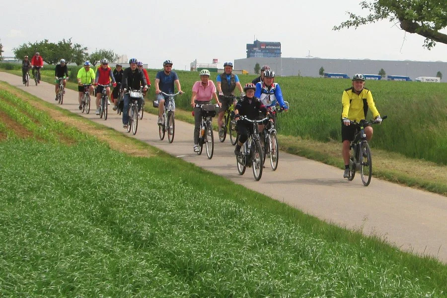 Gruppe von Fahrradfahrern auf einem Radweg in einer grünen Landschaft bei schönem Wetter.