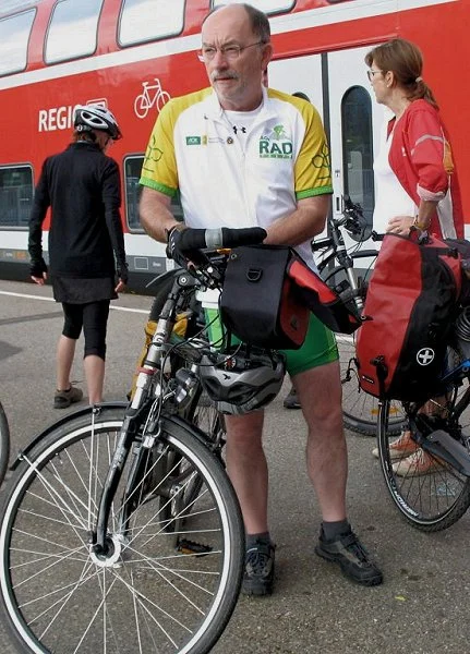 Mann in Radfahrer-Kleidung steht neben einem Mountainbike, im Hintergrund sind weitere Radfahrer und ein Bus.