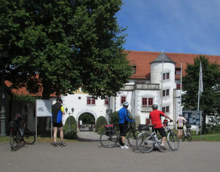 Fünf Radfahrer vor einem alten weißen Gebäude mit rotem Dach und Türmen in einem Park. Manche stehen neben ihren Fahrrädern, andere sind auf Fahrrädern und schauen sich das Gebäude an.
