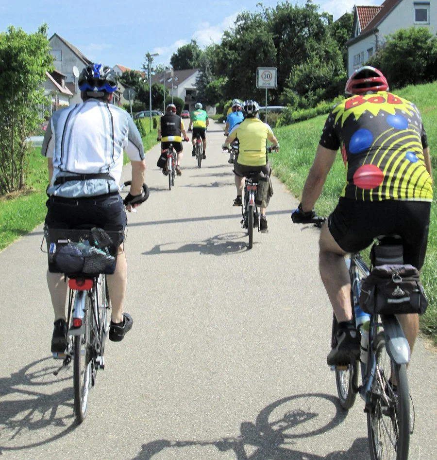 Mehrere Fahrradfahrer auf einem Weg in einer Wohngegend, einige tragen Helme und sportliche Kleidung, bei sonnigem Wetter.