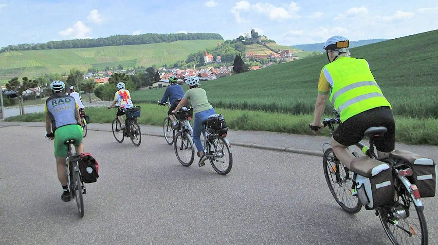 Sechs Fahrradfahrer, alle mit Helm, fahren auf einer Landstraße durch eine grüne Landschaft mit Hügeln und einem kleinen Dorf im Hintergrund.