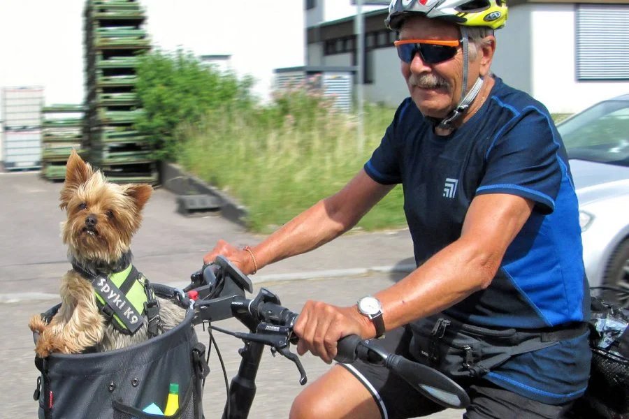 Ein Mann fährt Fahrrad mit einem Hund in einer Fahrradkörbe. Der Mann trägt einen Fahrradhelm und Sonnenbrille, der Hund trägt ein Sicherheitsgeschirr.