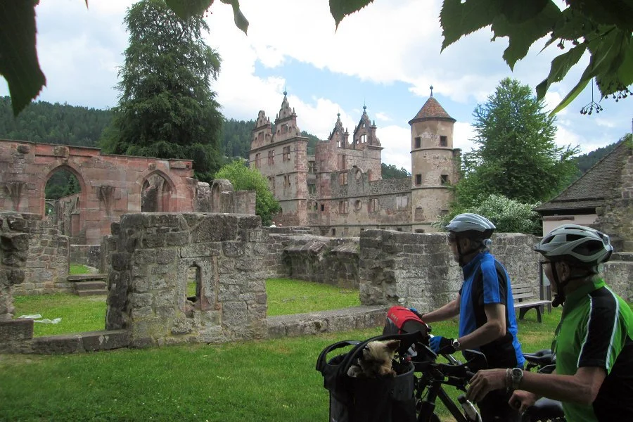 Zwei Radfahrer in Begleitung eines kleinen Hundes stehen vor den Ruinen eines alten Schlosses, im Hintergrund ein mittelalterliches Schloss mit Türmen, umgeben von Bäumen und Bergen.