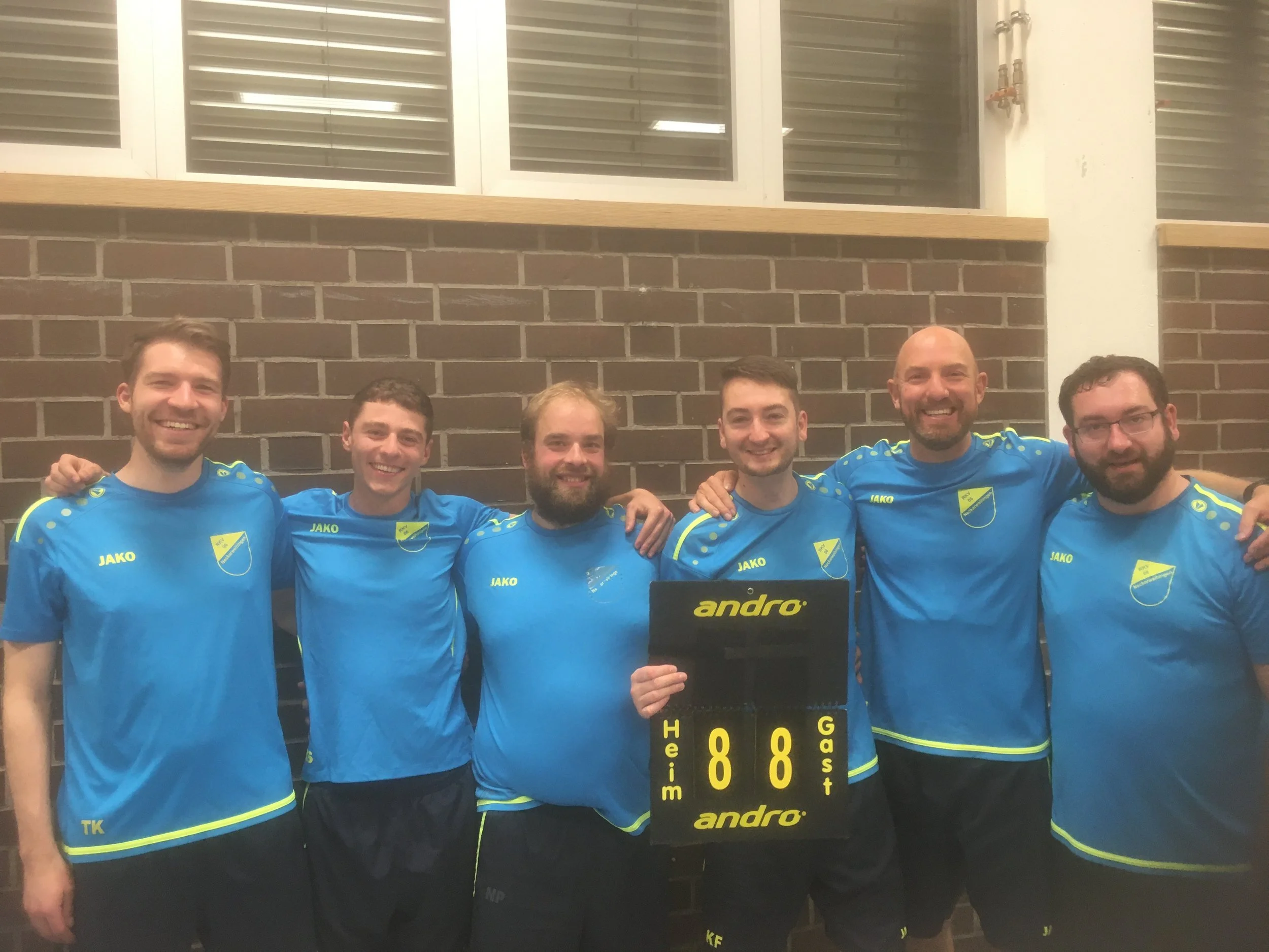Sieben Männer in blauen Sporttrikots stehen in einer Reihe vor einer Ziegelwand und halten ein Punktestück mit der Aufschrift 8-8. Sie lachen und haben die Arme um die Schultern des jeweils nebenstehenden.