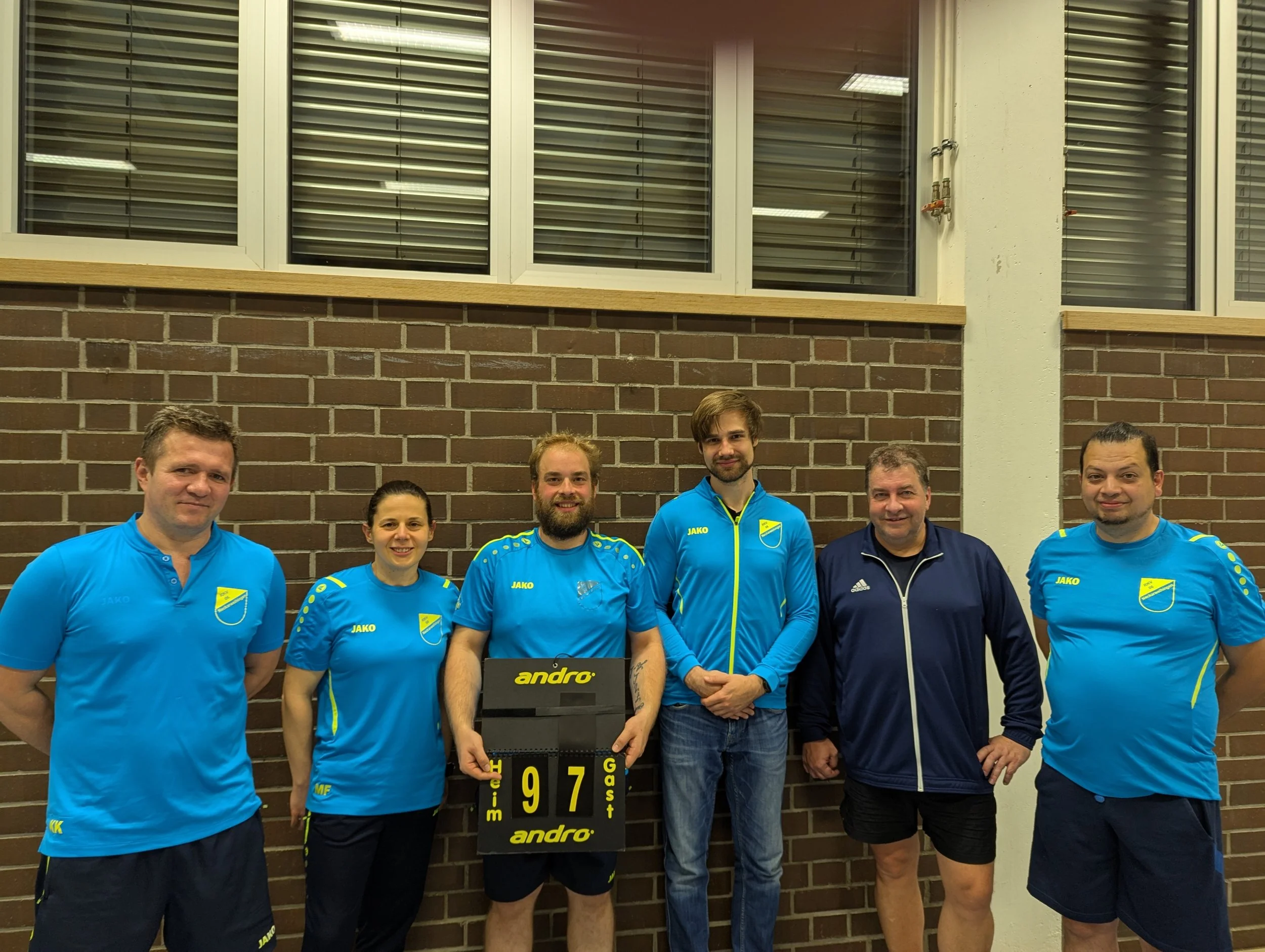 Gruppe von sechs Personen in blauen Sportanzügen, die nach einem Volleyballspiel jubeln, vor einer Ziegelwand mit Score-Tafel.