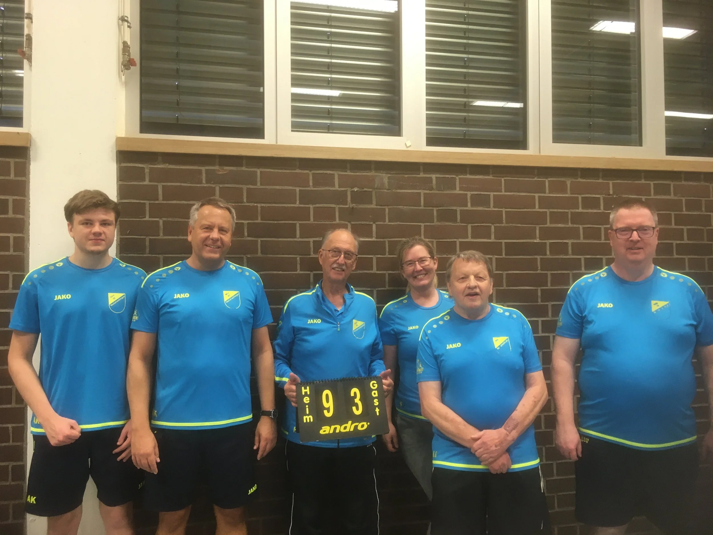 Gruppe von sechs Personen in blauen Sportoutfits, die eine ballspielende Mannschaft darstellen, vor einer Ziegelwand, mit einem Punktestandsschild.
