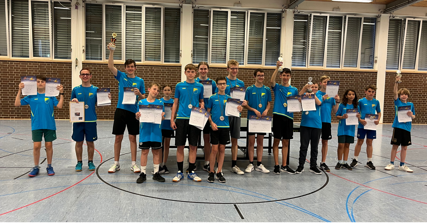 Jugendmannschaft in blauen Trikots bei einer Siegerehrung in einer Turnhalle, die Medaillen und Urkunden halten und Trophäen hochheben.