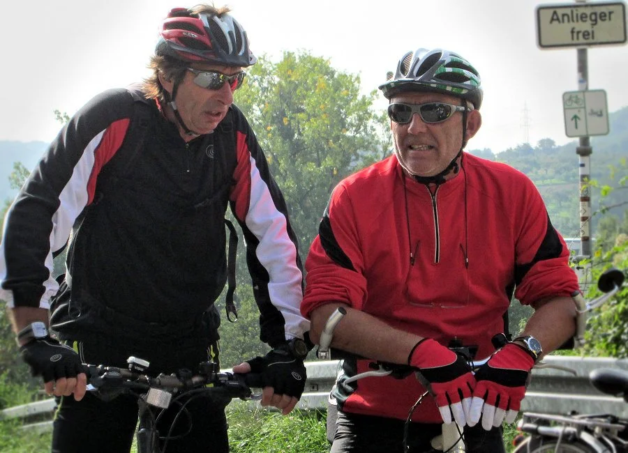 Zwei Mountainbiker in Fahrradkleidung mit Helmen und Sonnenbrillen stehen am Straßenrand. Einer trägt ein schwarzes Outfit, der andere ein rotes. Der Mann in Rot zeigt eine angespannte Mimik.