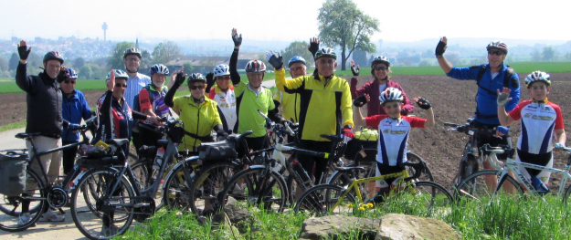 Gruppe von Radfahrern auf einer Wiese, die winken, mit Fahrradfahren Kleidung und Helm, im Hintergrund eine Feldlandschaft und eine Stadt am Horizont.