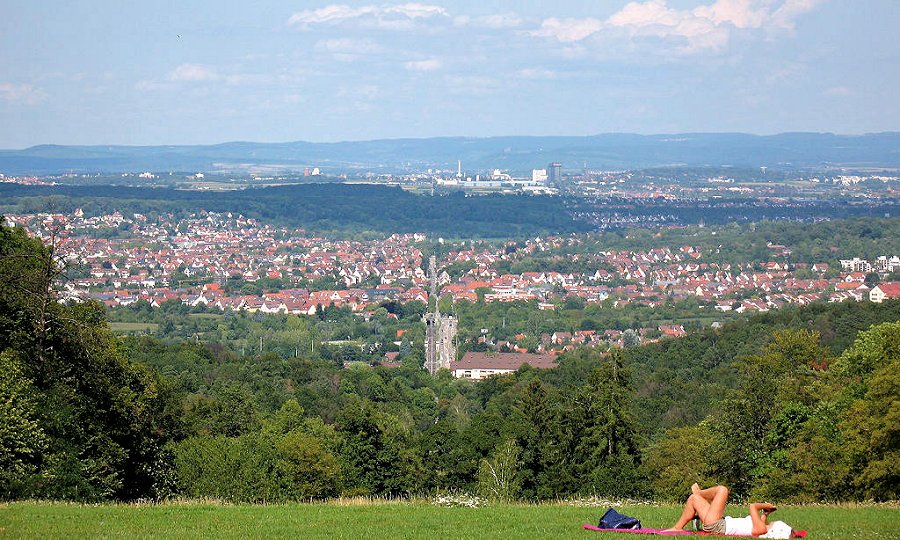 Blick auf eine Stadt mit Häusern und Industrie im Hintergrund, umgeben von grünen Wäldern, mit einer Person im Vordergrund, die auf einer Decke liegt und entspannt.