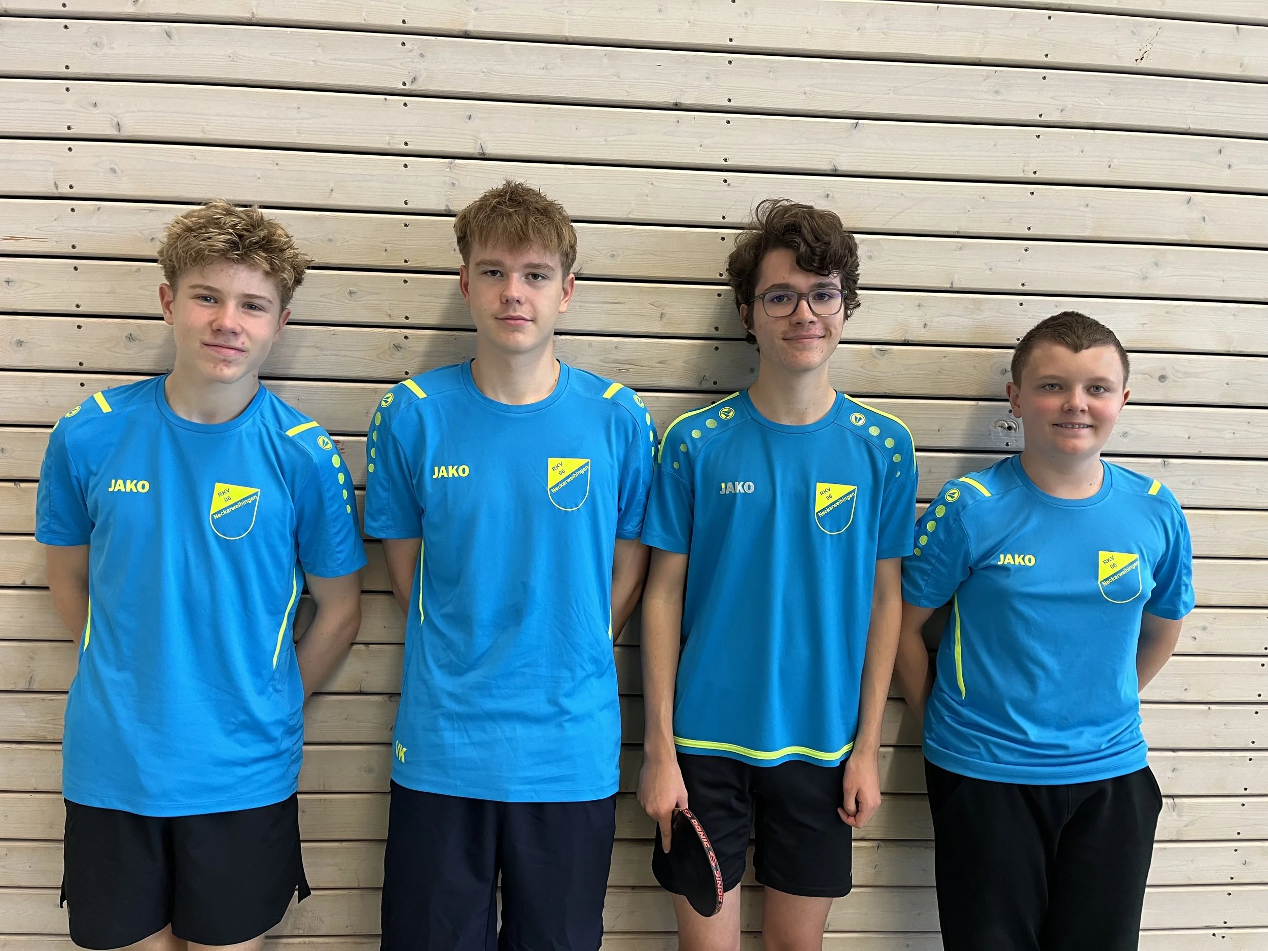 Vier junge Männer in blauen Sportt-Shirts vor einer Holzwand, alle stehen nebeneinander und schauen in die Kamera.