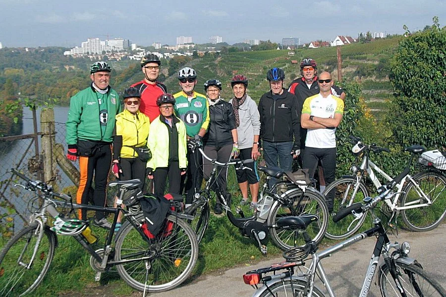 Gruppe von Radfahrern mit Fahrrädern auf einer Wiese neben einem Fluss, im Hintergrund Weinberge und Stadtgebäude, sonniger Tag.