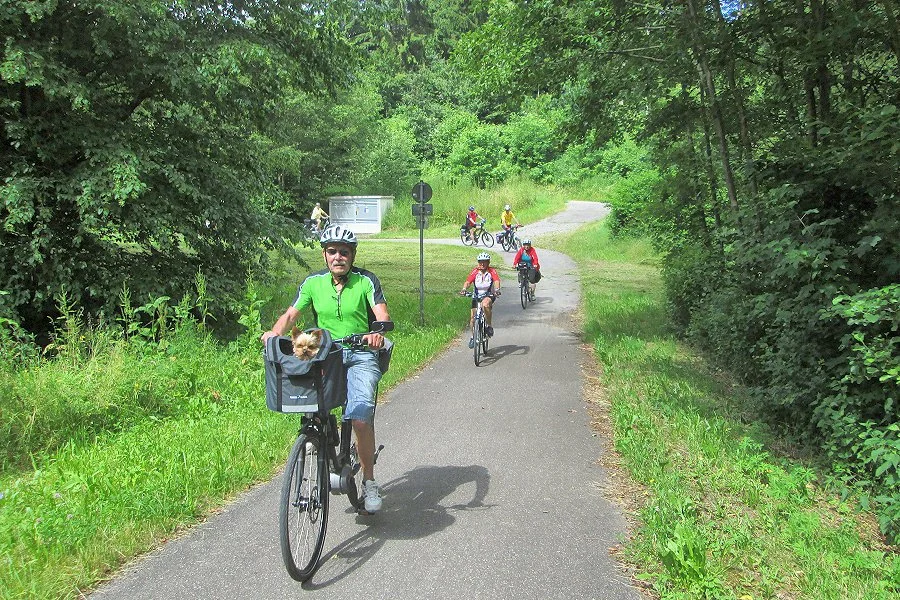 Gruppe von Radfahrern, einige Kinder, unterwegs in einem grünen Waldgebiet, wobei der vorderste Mann ein Fahrrad mit einem Hund in der Lenkertasche fährt.