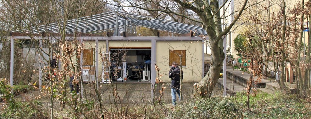 Ein Garten mit Bäumen und eine kleine, modern gestaltete Hütte. Vor der Hütte stehen zwei Menschen, einer fotografiert oder filmt die Szenerie, der andere ist in der Nähe des Eingangs.