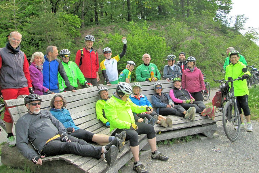 Gruppe von Radfahrern und Radfahrerinnen mit Helmen, sitzend und stehend auf einer Bank in einem grünen, bewaldeten Outdoor-Bereich, einige lächelnd in die Kamera schauend.