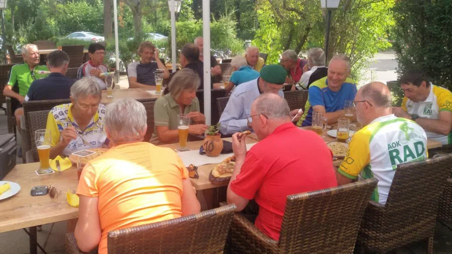 Eine Gruppe von Menschen sitzt an einem langen Tisch im Freien und genießt Essen und Getränke.