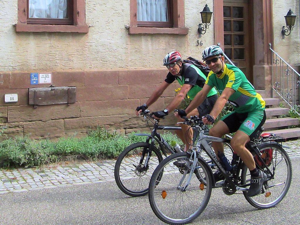 Zwei Männer in Radkleidung und Helmen fahren mit Fahrrädern auf einem gepflasterten Weg neben einem Haus mit schweren Wänden und Fenstern. Sie tragen Sonnenbrillen und lächeln.
