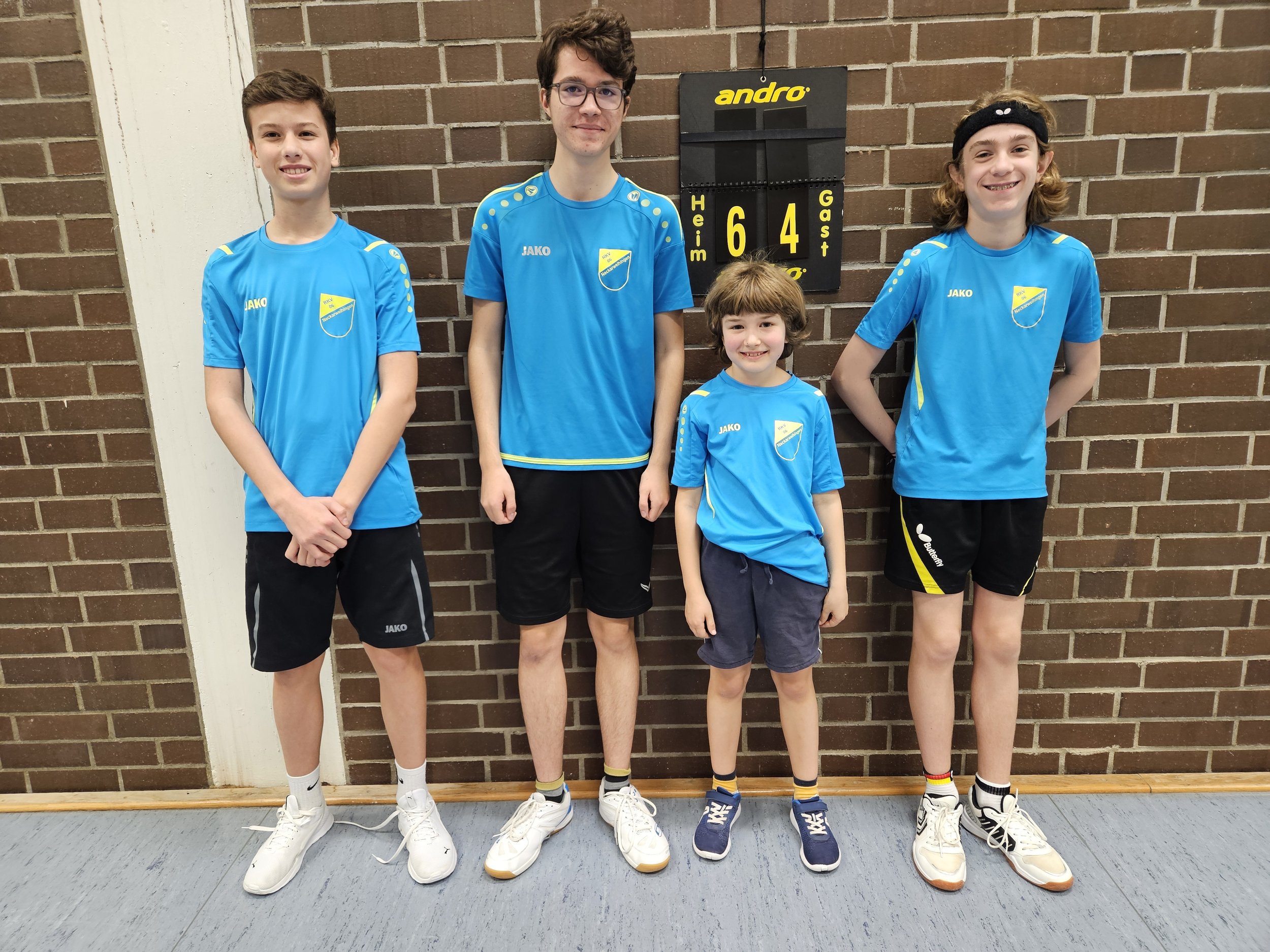 Vier Jugendliche in blauen Sporttrikots stehen vor einer Ziegelwand, neben einem Turnierscoreboard, das die Ergebnisse 6 zu 4 anzeigt.