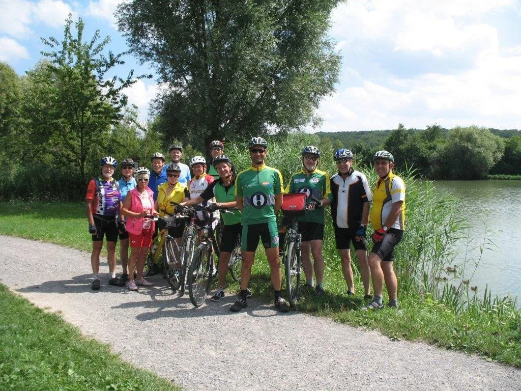 Eine Gruppe von Fahrradfahrern steht am Ufer eines Flusses unter Bäumen an einem sonnigen Tag.