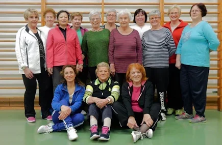 Gruppe älterer Frauen in einem Fitnessraum, einige stehen, zwei sitzen auf dem Boden, alle lachen und posieren für die Kamera.
