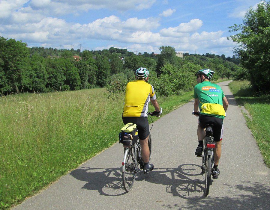 Zwei Fahrradfahrer in sportlicher Kleidung und Helmen fahren auf einem asphaltierten Weg durch eine grüne Landschaft mit Bäumen und Wiesen unter einem bewölkten Himmel.