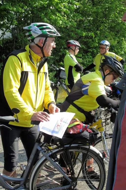 Eine Gruppe von Radfahrern in leuchtend gelben Jacken, mit Fahrradhelmen, bereitet sich auf eine Tour vor. Ein Mann liest eine Karte oder ein Blatt, während er auf einem Fahrrad sitzt, neben anderen Radfahrern, die ihre Fahrräder bereitstellen.