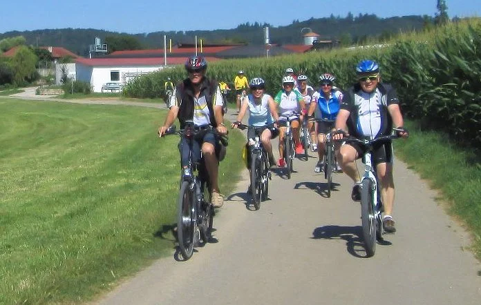 Gruppe von acht Personen, die Fahrrad fahren, auf einem asphaltierten Weg in einer ländlichen Gegend mit Häusern, Gärten und bunten Feldern im Hintergrund, sonniges Wetter.