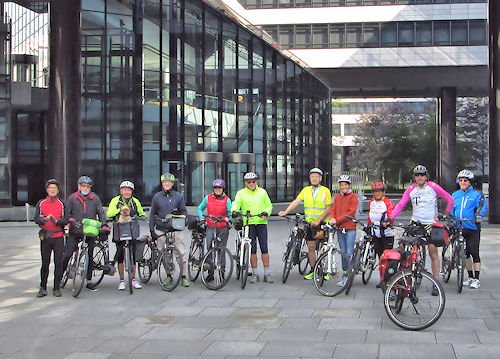 Gruppe von zehn Radfahrern, die vor einem modernen Glasgebäude mit mehreren Stockwerken stehen.