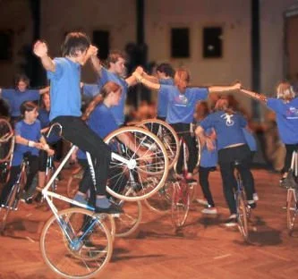 Kinder in blauen T-Shirts üben im Kreis auf Rollstühlen und einem Fahrrad, auf einer Bühne im Freien.