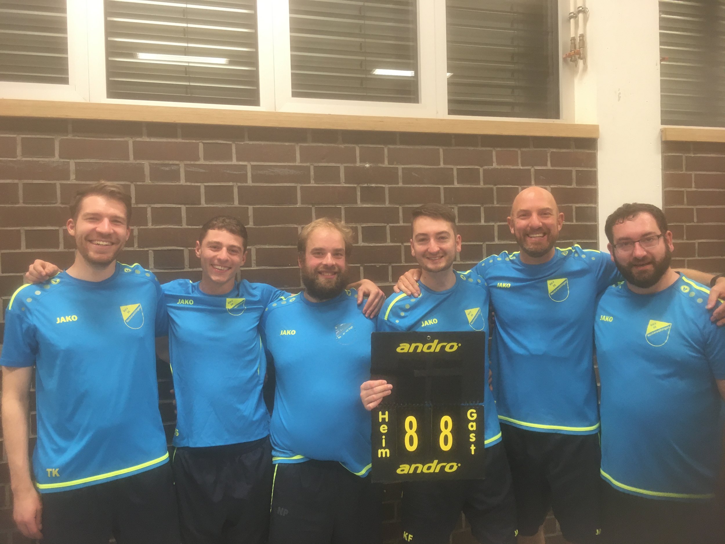 Eine Gruppe von sieben Männern in blauen Sporttrikots, die um ein Tischkicker-Board mit der Aufschrift 'andro' steht, in einer indoor Sporthalle vor einer Ziegelwand.