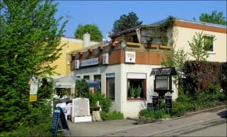 Ein zweistöckiges Gebäude mit einem Café im Erdgeschoss, umgeben von Bäumen und Pflanzen, bei sonnigem Wetter.