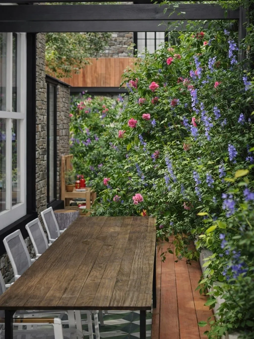 Planting-led garden with lush climbing plants and layered borders surrounding a timber dining table, set against stone walls and contemporary architectural details.