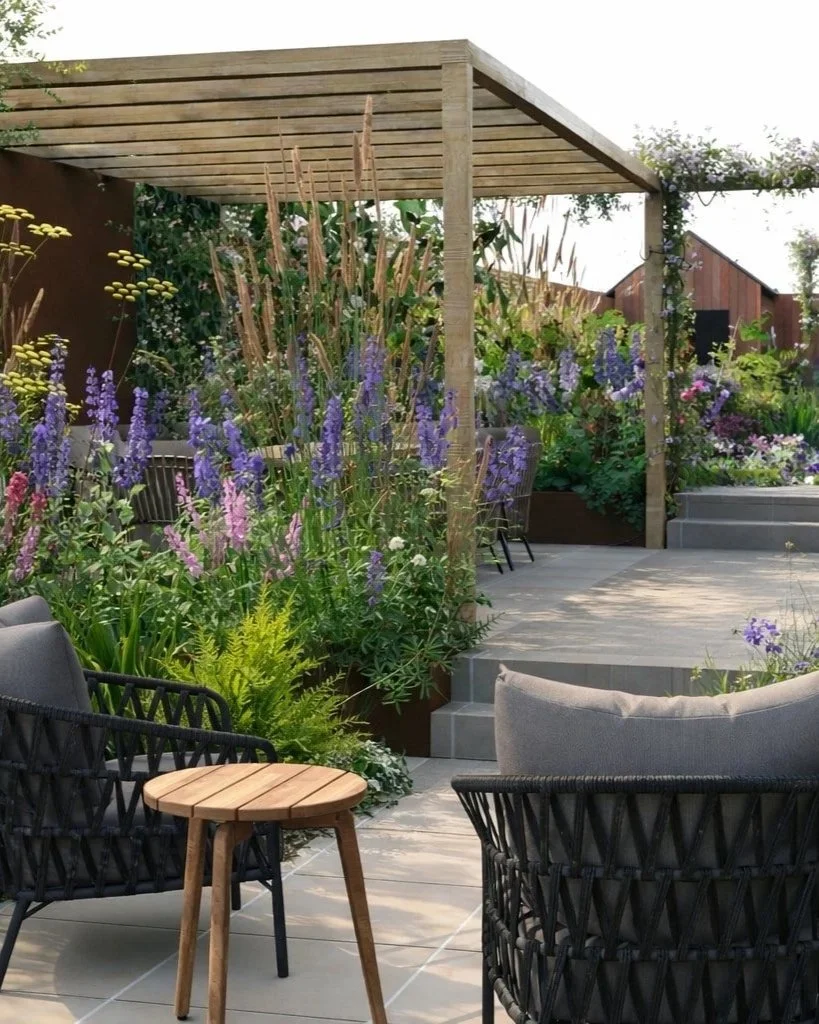 A render of close up of a garden design showing wooden pergola, 8 seater dining table, 2 chairs and coffee table and lots of colourful perennial planting.