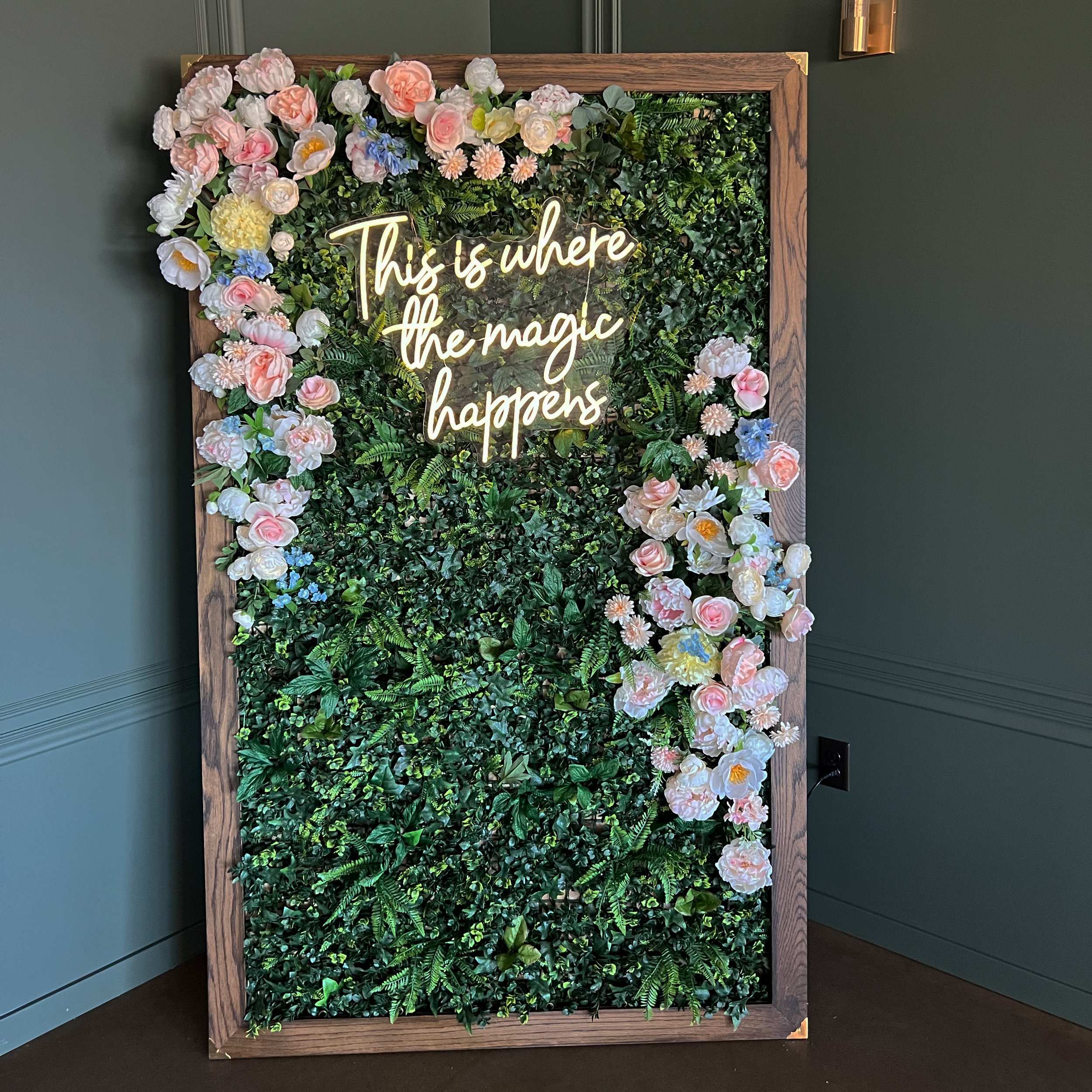 image of a frame with greenery as the backdrop with an LED sign that says, "This is where the magic happens"