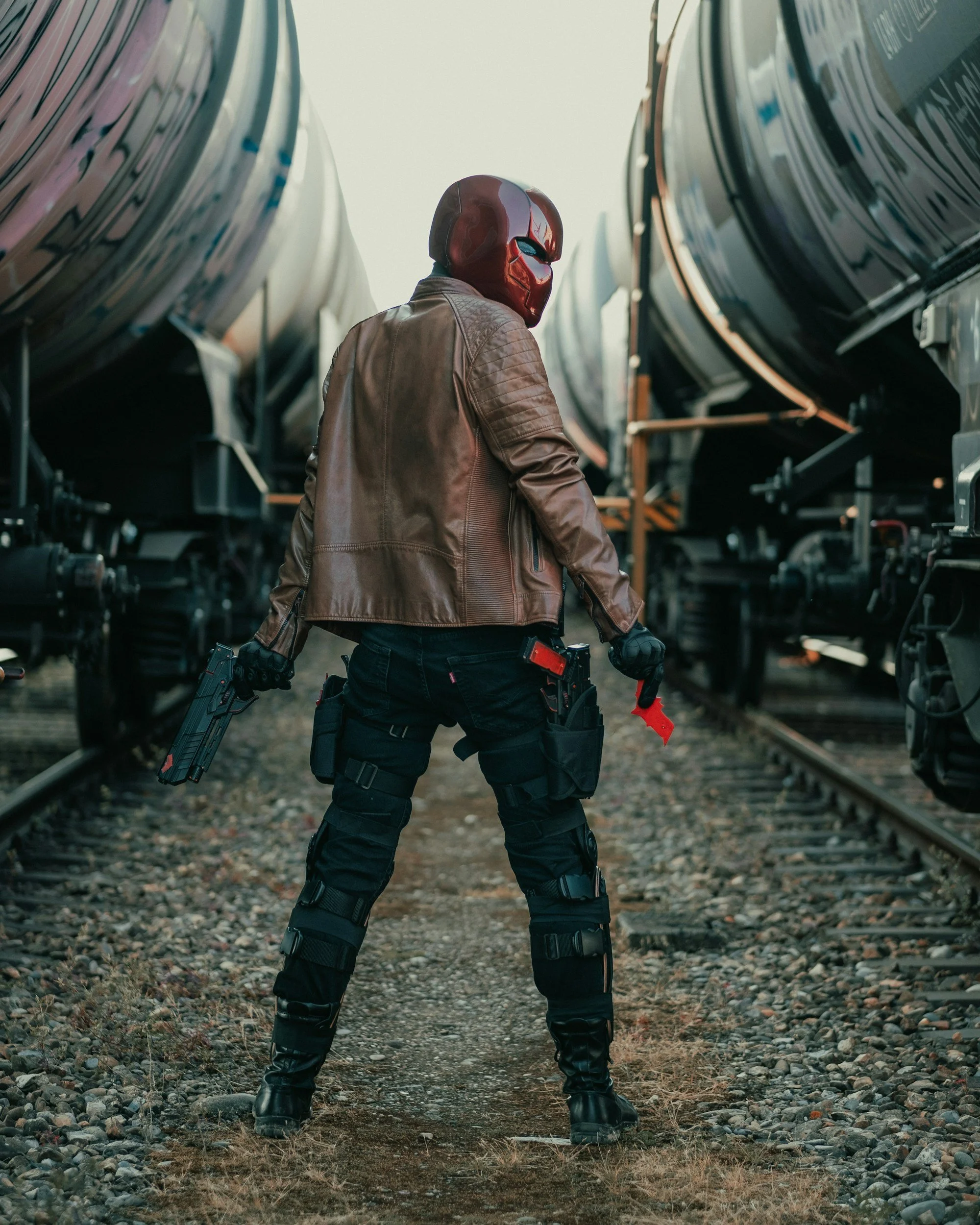 A person wearing a red helmet, brown leather jacket, and tactical black pants standing between two train cars on a railroad track, holding a gun in each hand.