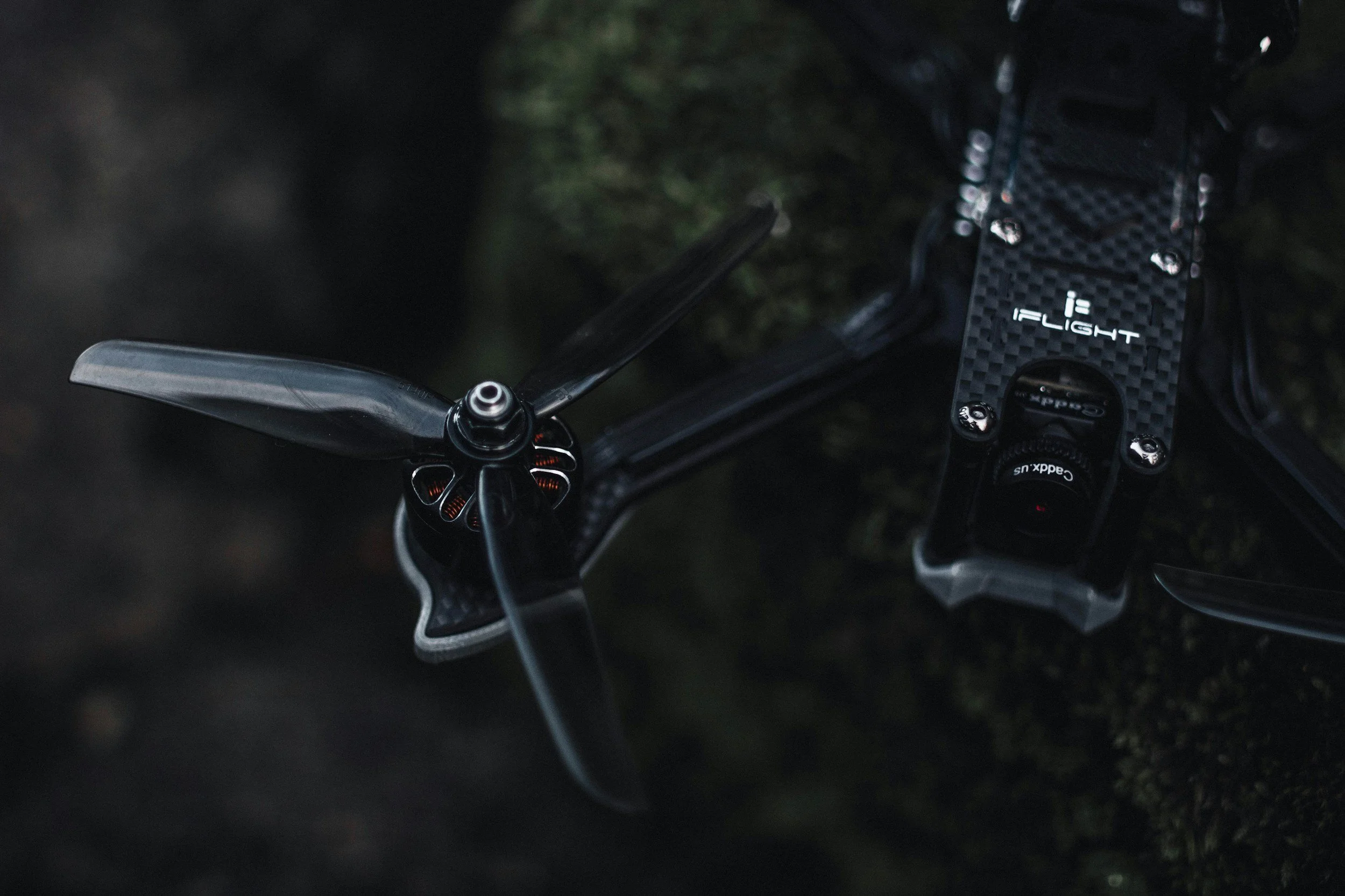 A close-up of a black drone with a carbon fiber top and a camera, resting on a mossy ground in a forest. The drone has three propellers and the camera is labeled 'GoPro'.