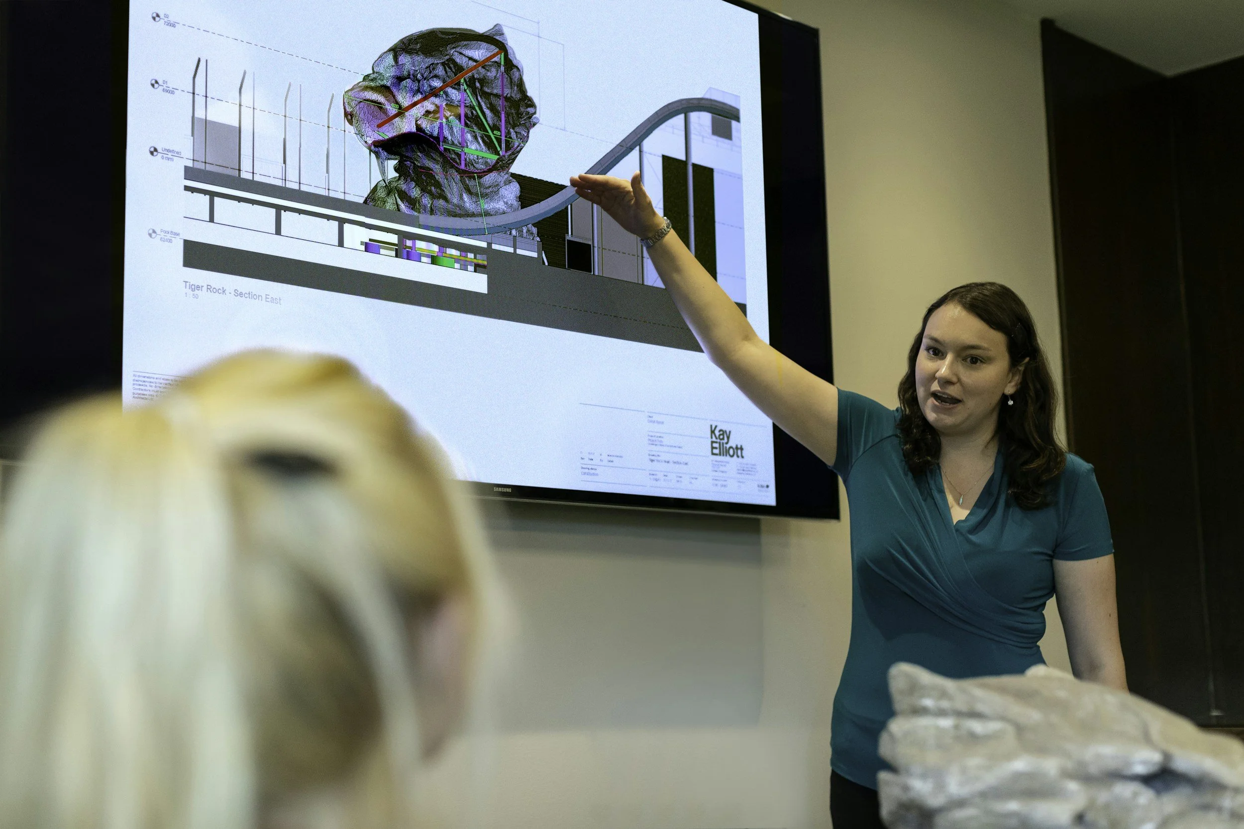 A woman giving a presentation to a small audience in a conference room. She is pointing at a large screen displaying a 3D geological model or diagram and some technical data.