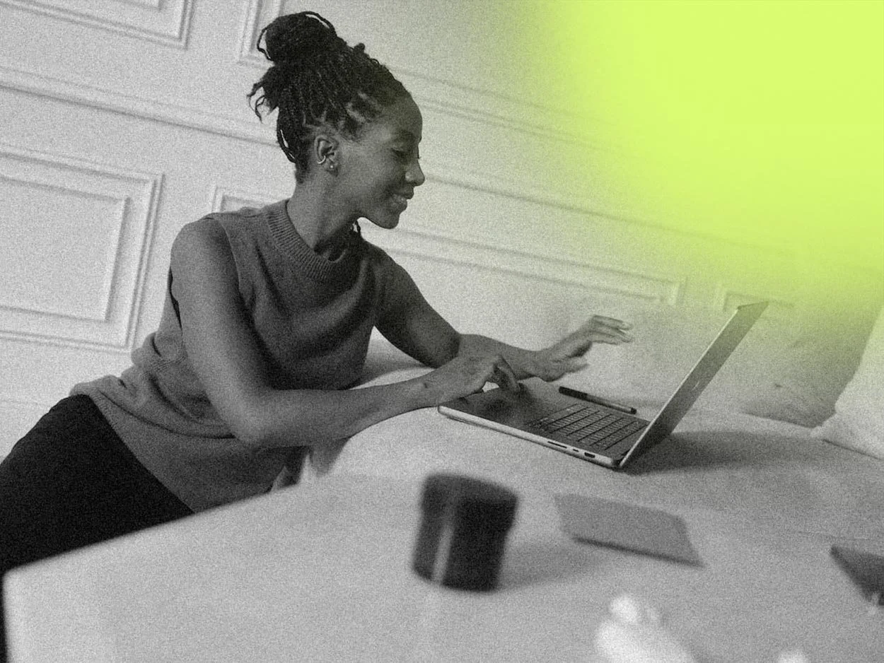 A woman with dreadlocks in a bun working on a laptop at a table, smiling with a relaxed posture, in black and white with a yellow highlight in the top right corner.