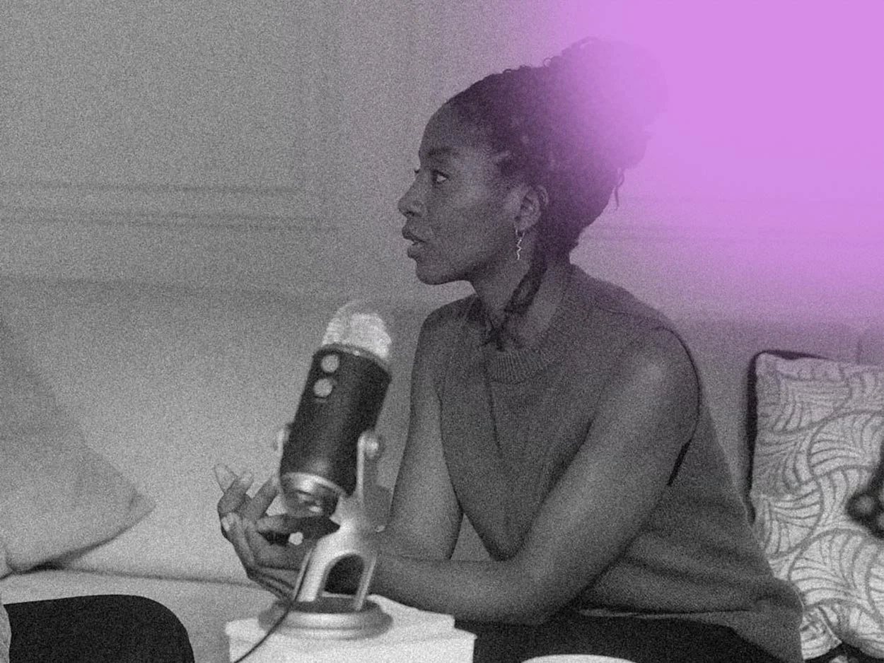 A black and white photo of a woman with braided hair, sitting and speaking into a microphone on a table, with a decorative pillow behind her.