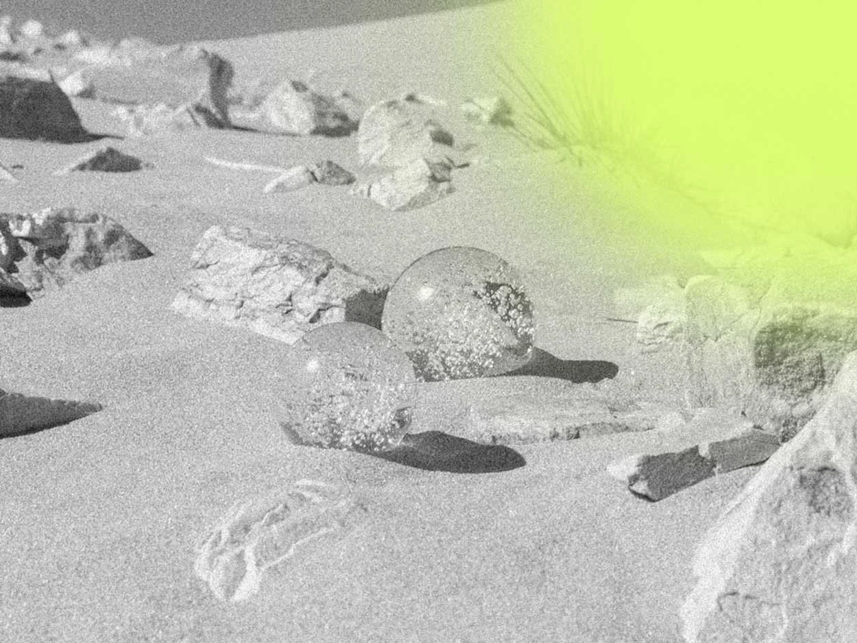 Close-up of a sandy terrain with rocks and pebbles, two glass marbles with reflections, and a blurred palm leaf in the yellowish sunlight.