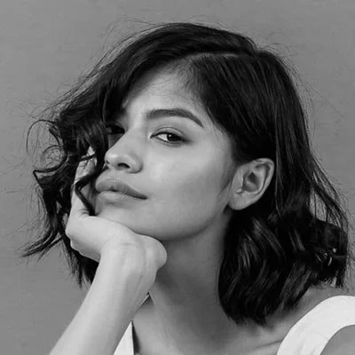 Black and white portrait of a woman with short wavy hair, resting her chin on her hand and looking at the camera with a slight smile.