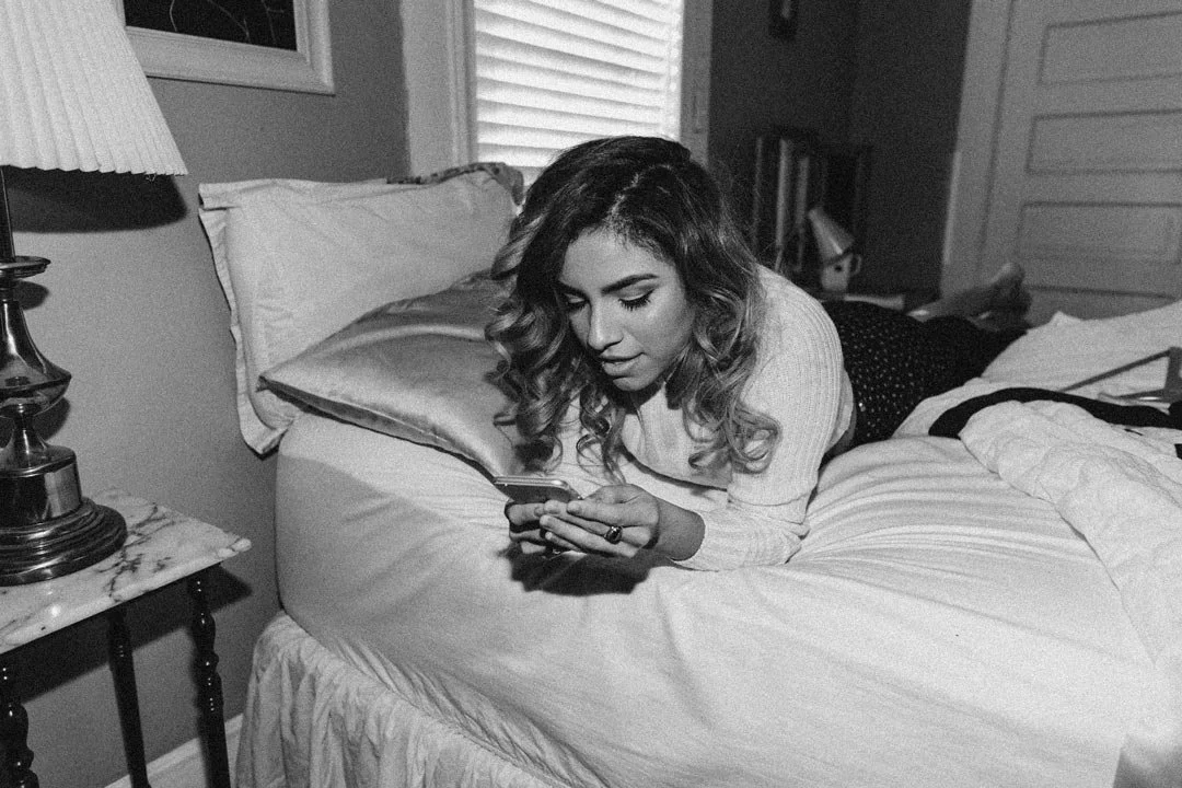 A woman with wavy hair lying on her stomach on a bed, looking at her phone in a bedroom with a nightstand, lamp, and window.