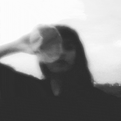 A person with long dark hair covering their face, wearing a dark top, and holding their hand to their head in a contemplative pose.